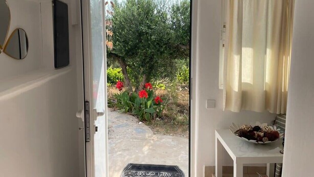 An open door leading to a garden with red flowers and a white table with a bowl of fruit. - Gorgeous White Marsis Apartment in Paros (Paros)