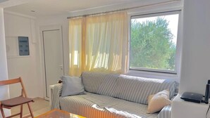 A living room with a sofa, a glass coffee table, a chair, and a window with curtains. - Gorgeous White Marsis Apartment in Paros (Paros)