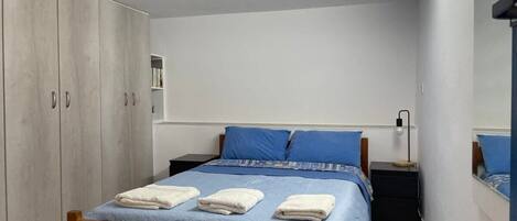 Un dormitorio con cama de madera, ropa de cama azul, una mesita de noche, un armario y un espejo en la pared.