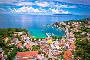An aerial view of a coastal town with a marina, buildings with red roofs, and a clear blue sea.