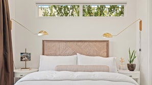 Une chambre à coucher avec un lit, deux lampes de chevet, une table de nuit, un tableau encadré, une plante en pot et une fenêtre donnant sur des arbres.