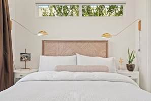 Een slaapkamer met een bed, twee bedlampen, een nachtkastje, een lijst met een afbeelding, een potplant en een raam met uitzicht op bomen.