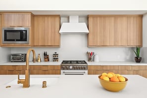 A modern kitchen with wooden cabinets, a stainless steel microwave, a gas stove, a built-in oven, a sink with a gold faucet, and a bowl of fruit on the counter.