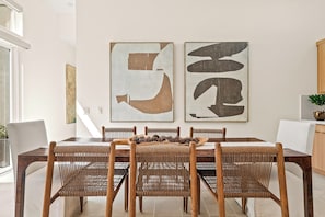 A dining area with a wooden table and woven chairs, a white wall with abstract art, and a potted plant on a cabinet.