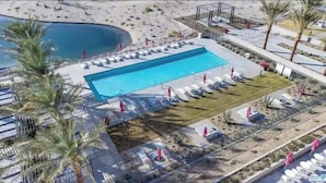 Vue aérienne d’un espace piscine avec des chaises longues et des parasols, entouré de palmiers et d’un paysage sablonneux.