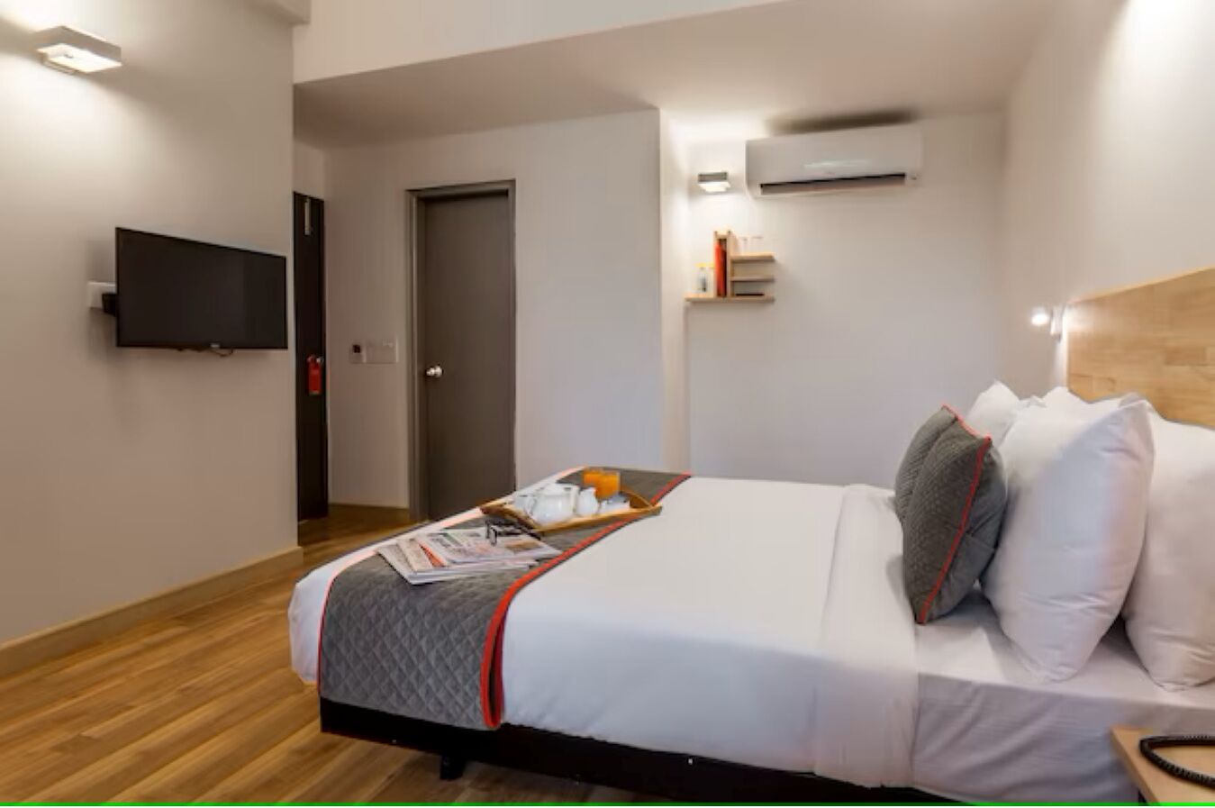 A hotel room with a bed, a flat-screen TV mounted on the wall, an air conditioning unit, and a small shelf with books and bottles.