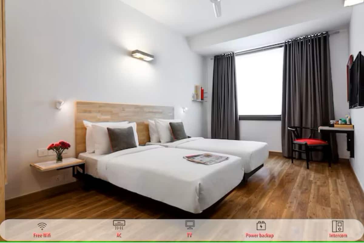 A hotel room with two beds, a desk, a chair, a window with curtains, and a wall-mounted light.