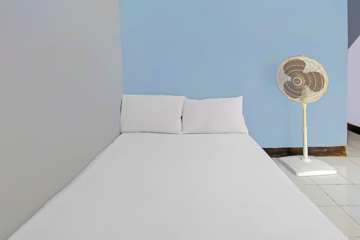 A bed with white bedding, a standing fan, and a blue wall.