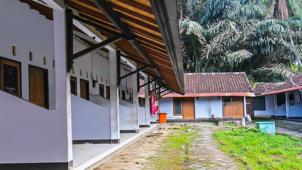 A covered walkway with a tiled floor, leading to a building with multiple windows and a red-tiled roof. - Hotel O Sukandi Homestay Near The Semeton Homestay (Janapria)