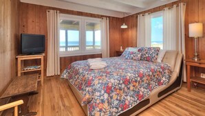 Un dormitorio con suelo de madera, cama con cubrecama floral, televisor sobre un mueble de madera y vista al mar a través de amplios ventanales.