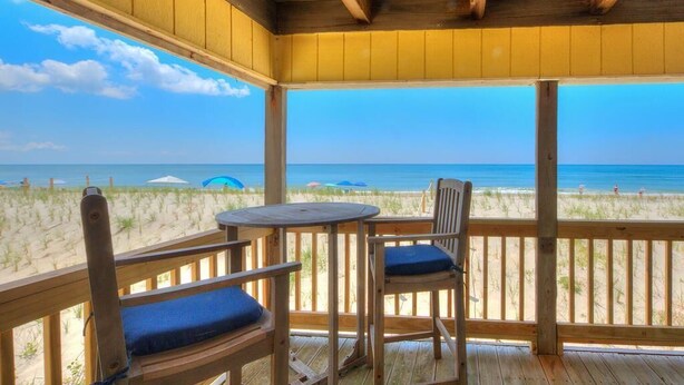 Una terraza de madera con dos sillas y una mesa, con vista a una playa con sombrillas y el océano.