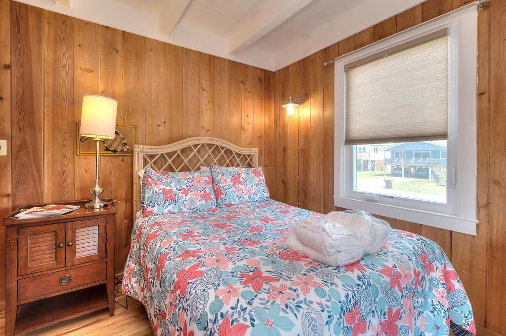 A bedroom with wooden paneling, a bed with floral bedding, a nightstand with a lamp, and a window with a view.