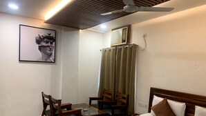 A hotel room with a bed, two chairs, a small table, a ceiling fan, an air conditioning unit, and a framed picture on the wall. - Hotel Avkaash  (Ambala)