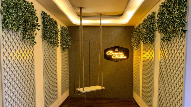A narrow hallway with a swing chair, decorative foliage, and a neon sign for Brannies Bistro. - Hotel Avkaash  (Ambala)
