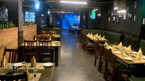 A restaurant with wooden tables and chairs, some booths, and a stage in the background. - Hotel Avkaash  (Ambala)