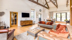 A spacious living room with a high ceiling, wooden beams, a television, a sofa, a coffee table, and a dining area with a view through large windows.