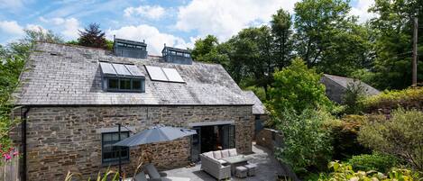 A stone house with a sloped roof, a patio with outdoor furniture, and a garden with various plants.