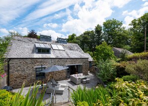 A stone house with a sloped roof, a patio with outdoor furniture, and a garden with various plants.