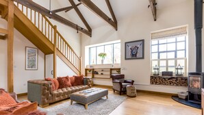 A spacious living room with a high ceiling, wooden beams, a staircase, a fireplace, and a large window.