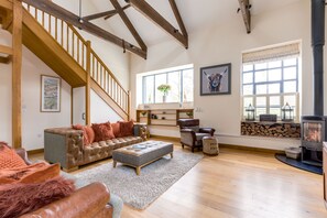 A spacious living room with a high ceiling, wooden beams, a staircase, a fireplace, and a large window.