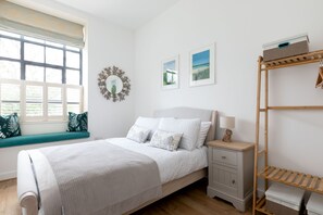 A bedroom with a bed, a bedside table, a lamp, a shelf, and a window with blinds.