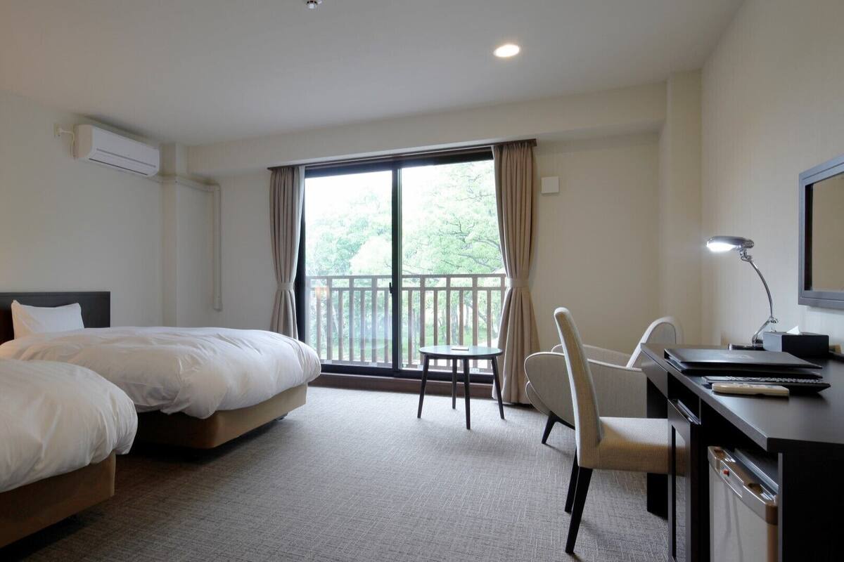 A hotel room with two beds, a desk with a chair, a small table, and a balcony with a view of trees.