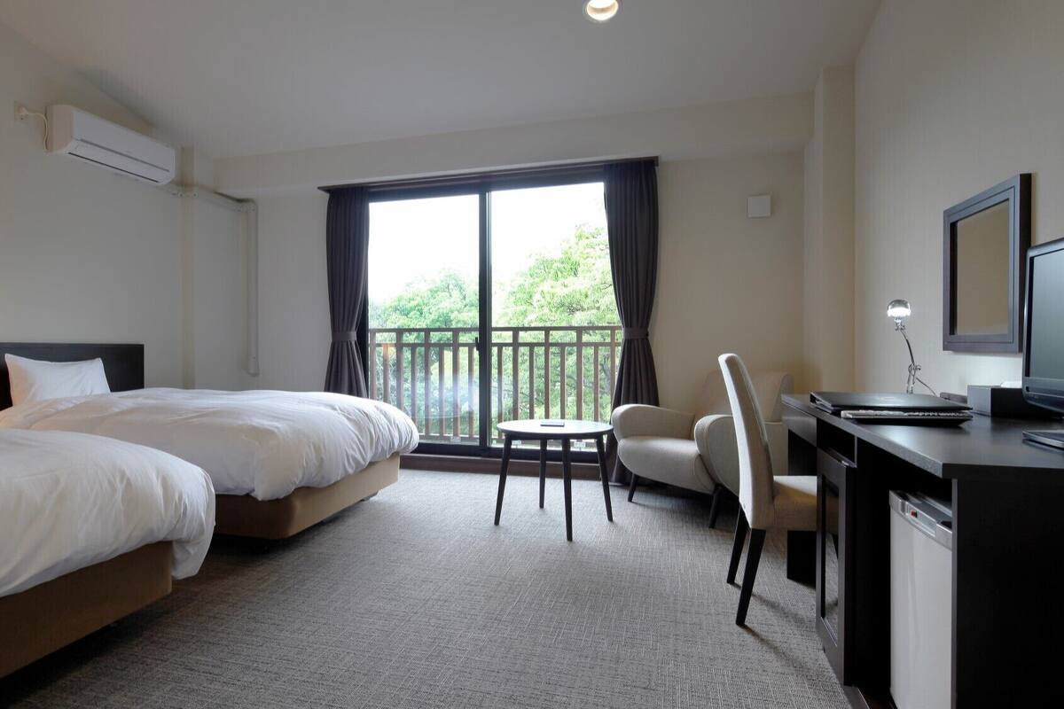 A hotel room with two beds, a desk with a computer, a chair, and a large window with a view of greenery.