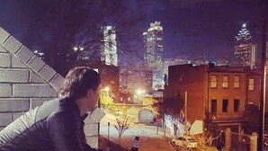 Una persona a gairebé mig cos, inclinada per una finestra amb vista a un paisatge urbà de nit.