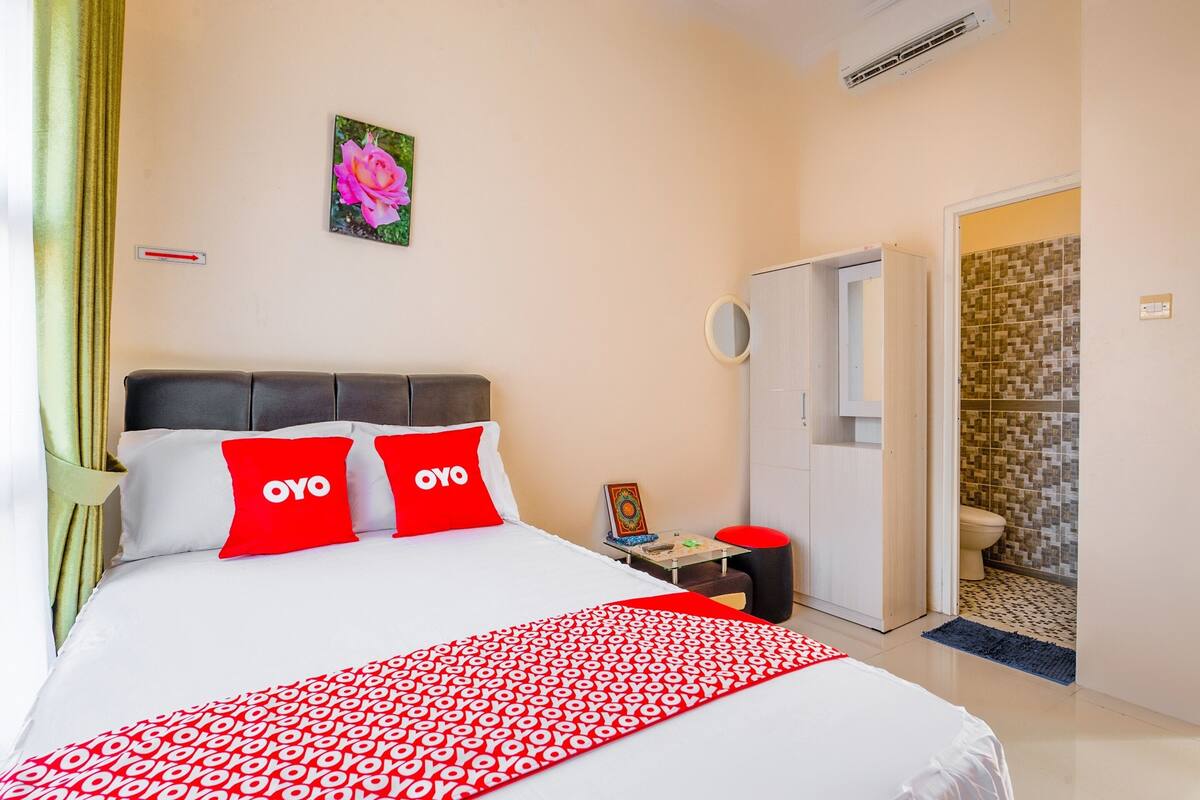 Une chambre d’hôtel avec un lit, une table de chevet, un miroir et une salle de bain visible à travers une porte ouverte.