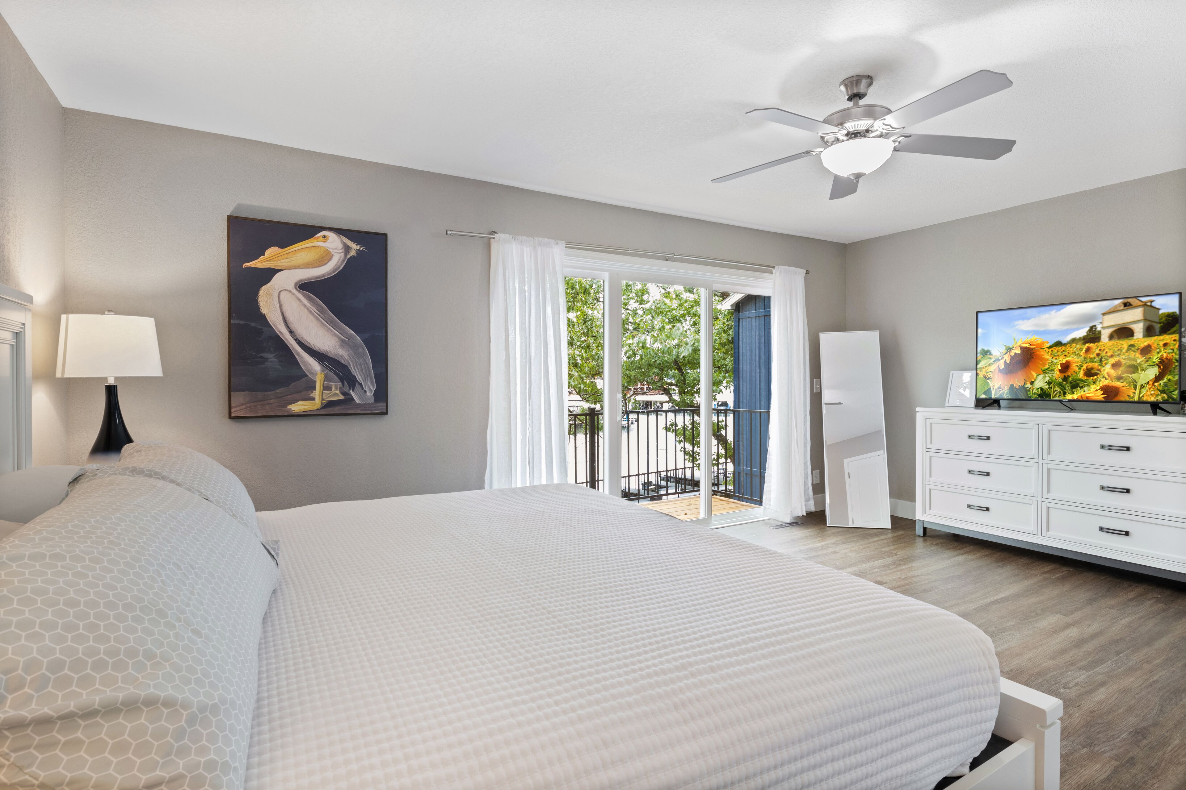 A bedroom with a large bed, a ceiling fan, a painting of a pelican, a television, and a view of the outdoors through a sliding door.