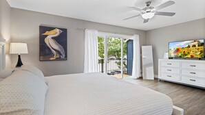 A bedroom with a large bed, a ceiling fan, a painting of a pelican, a television, and a view of the outdoors through a sliding door.