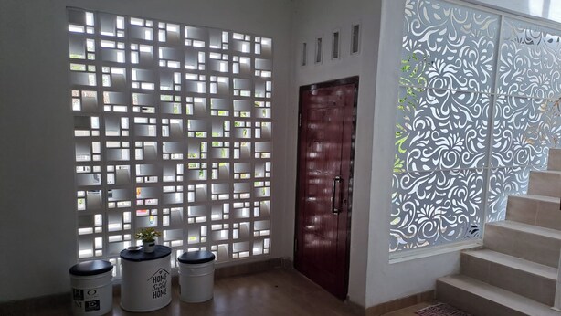 A room with a wooden floor, a red door, and a decorative white partition with cut-out patterns. There are two white containers with lids and a small potted plant on the floor. - Hotel O Safira Guest House Danau Ranau (Muaradua)