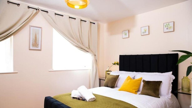 Un lit bien fait, recouvert d’une couverture verte, avec des oreillers blancs et une tête de lit noire. Deux tableaux encadrés sont accrochés au mur, il y a une fenêtre avec des rideaux et une plante posée sur une table d’appoint.