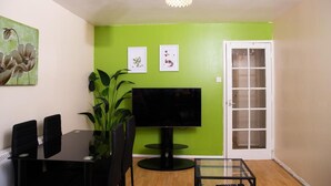 Un salon avec un mur vert, un téléviseur à écran plat, une table à manger avec des chaises et une plante en pot.