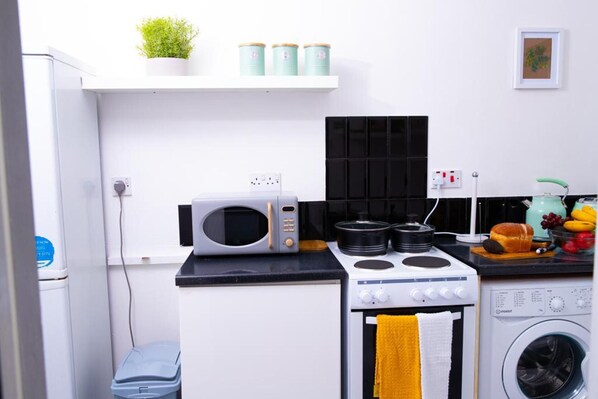 A compact kitchen with a microwave, stove, and washing machine. There are shelves with plants and containers, and a small trash bin. - Inviting 1-bed Apartment in London, Near Hotsput (London)