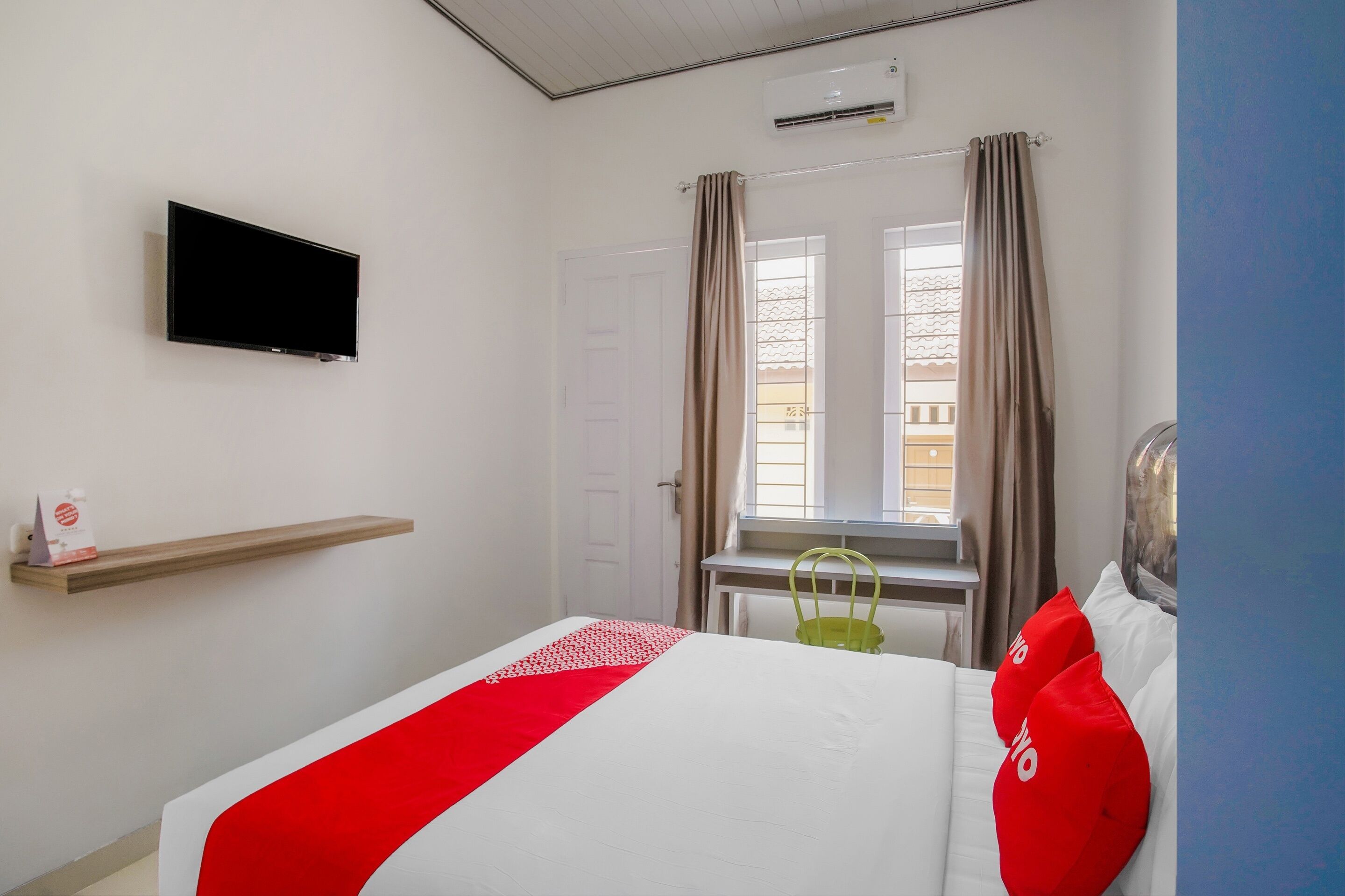 Een hotelkamer met een bed, een aan de muur bevestigde televisie, een raam met gordijnen en een klein tafeltje met een stoel.