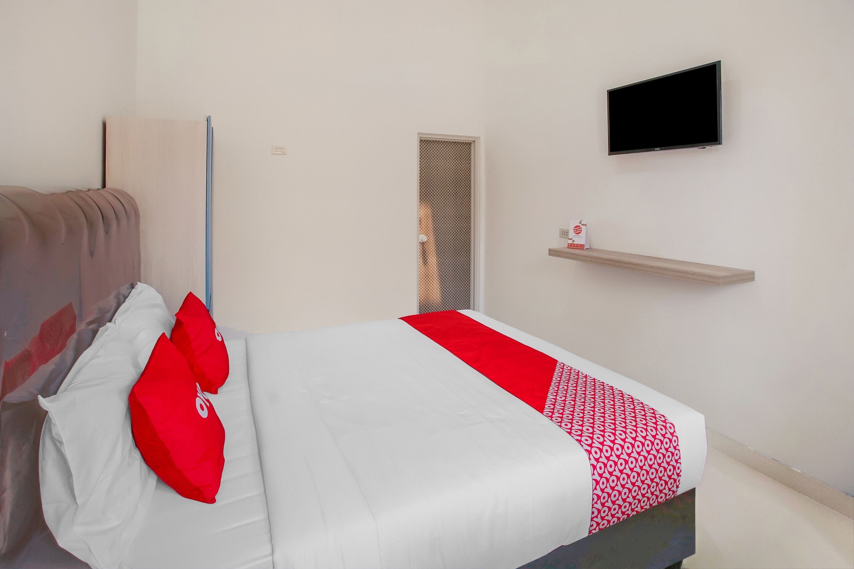 A neatly made bed with white and red bedding, a television mounted on the wall, and a small shelf with a decorative item.