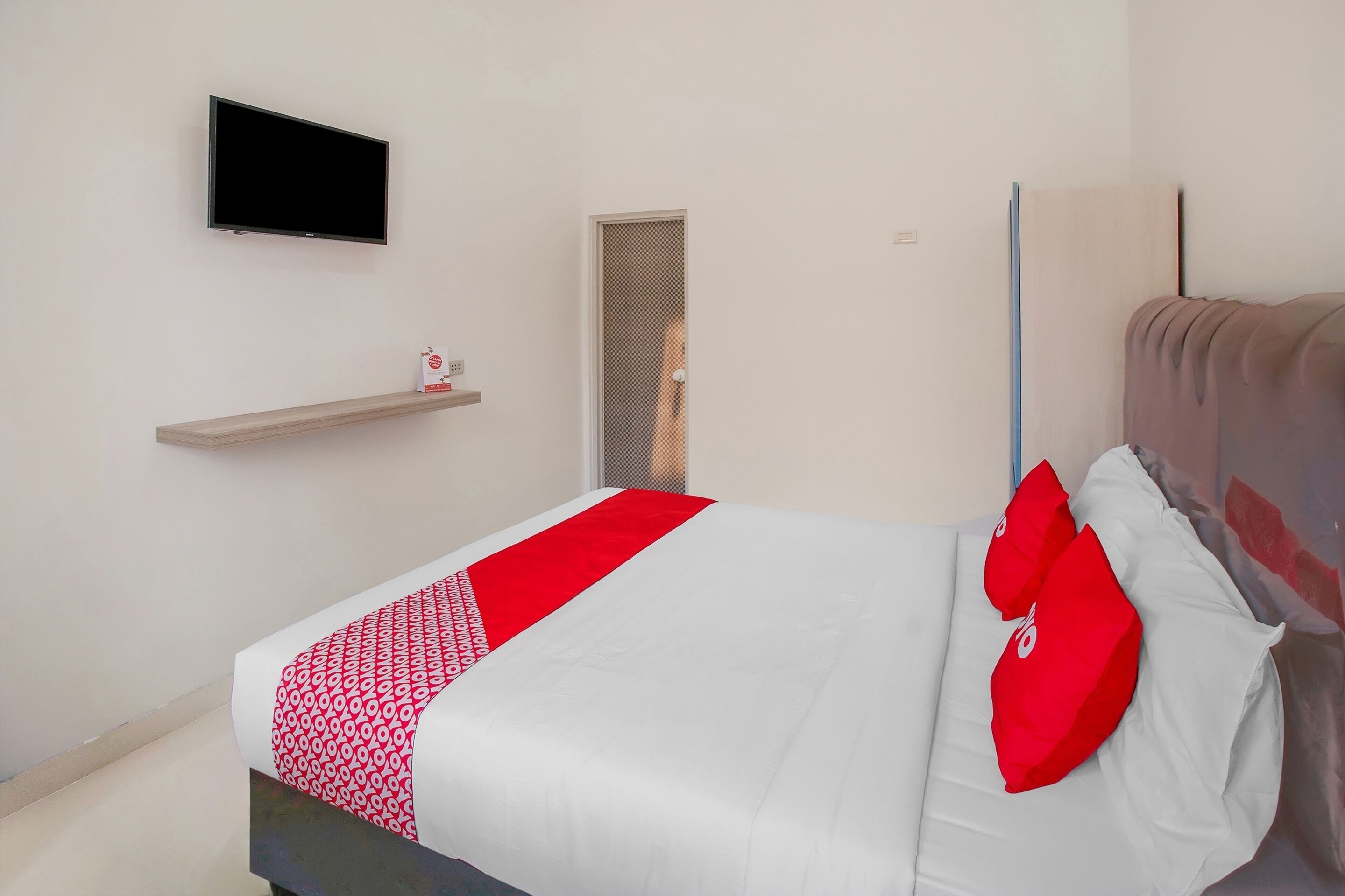A neatly made bed with a red and white bedspread, a television mounted on the wall, and a small shelf with a red object.