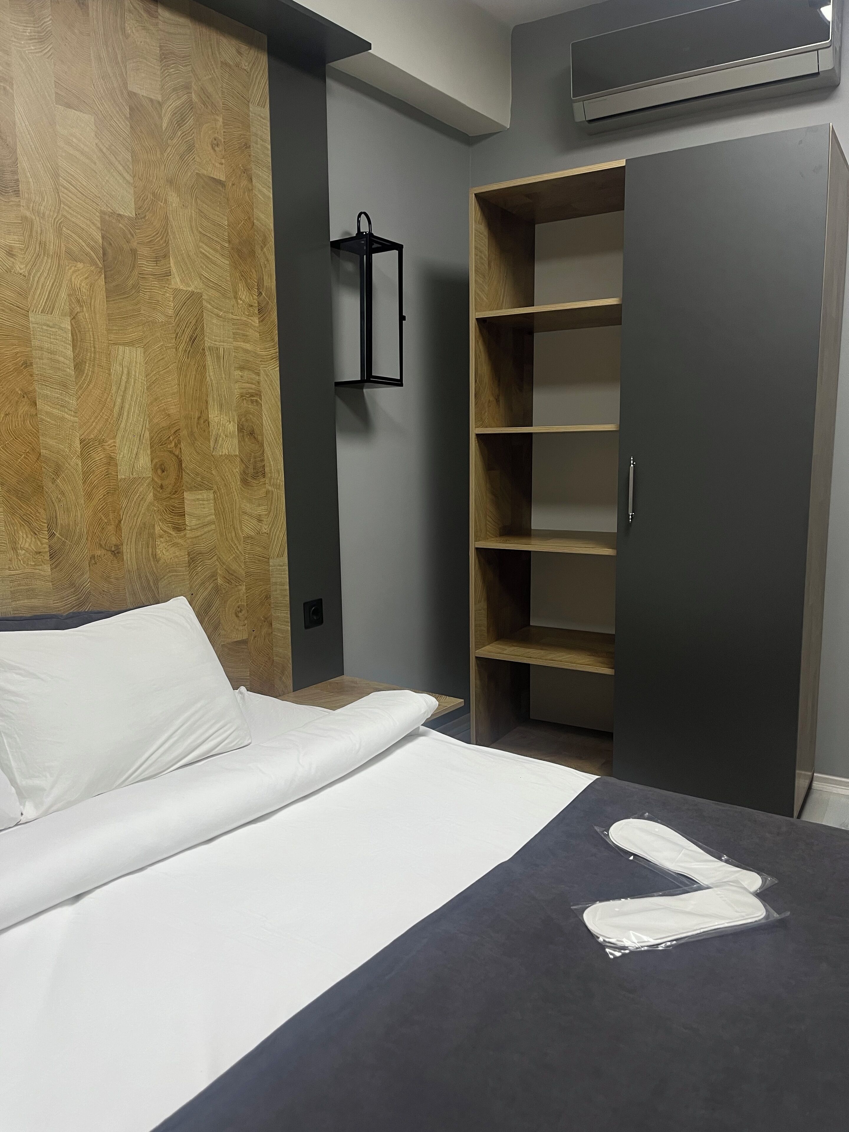 Quarto de hotel moderno com parede em madeira, cama com roupa de cama branca, guarda-roupa cinza e estante embutida.