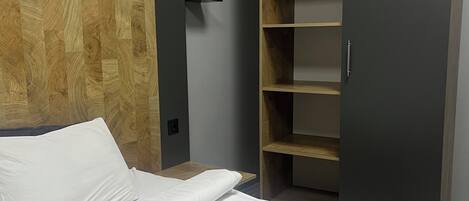 A modern hotel room with a wooden accent wall, a bed with white linens, a grey wardrobe, and a built-in shelf unit.