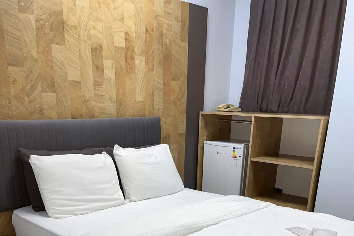 Un lit bien fait, avec une literie blanche, une tête de lit grise et une tête de lit en bois. On y voit une petite table de chevet en bois et une table de chevet blanche. Un mur blanc orné d’un panneau en bois est visible derrière le lit.
