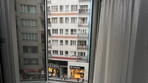Une vue depuis une fenêtre donnant sur un immeuble à plusieurs étages, doté de balcons et de devantures de magasins, notamment ceux des enseignes « AKARYA » et « Mathla ».