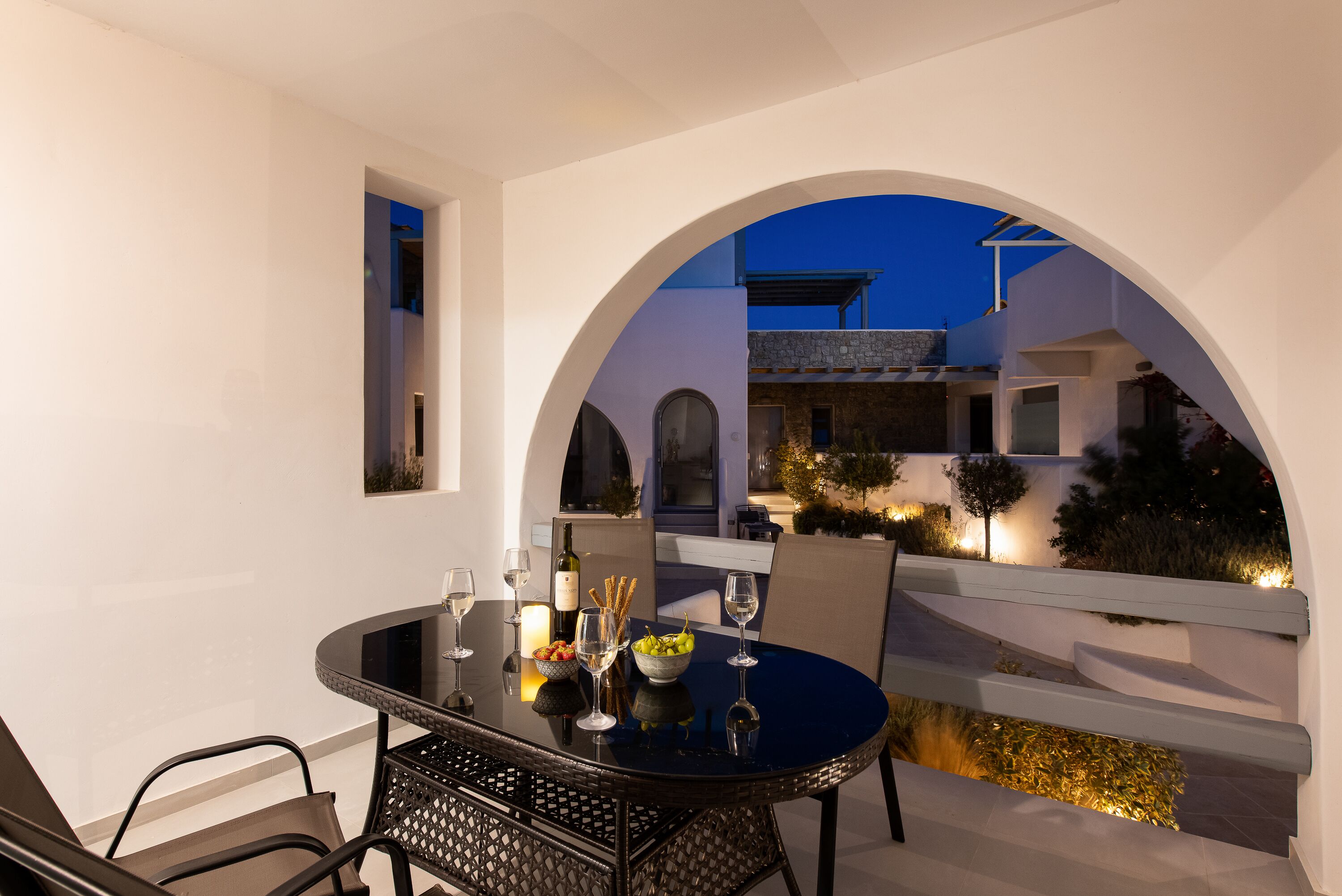 A dining area with a round table, chairs, and a view through an arched doorway to a building with stone walls and a blue sky.