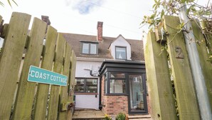 Une petite maison avec une porte en bois, une enseigne indiquant « COAST COTTAGE », et une maison en briques dotée d’une fenêtre à cadre noir.
