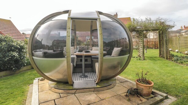 Une sorte de bulle sphérique en verre, avec une petite table et un espace salon à l’intérieur, installée sur une terrasse en pierre, avec un jardin et une pergola en bois en arrière-plan.