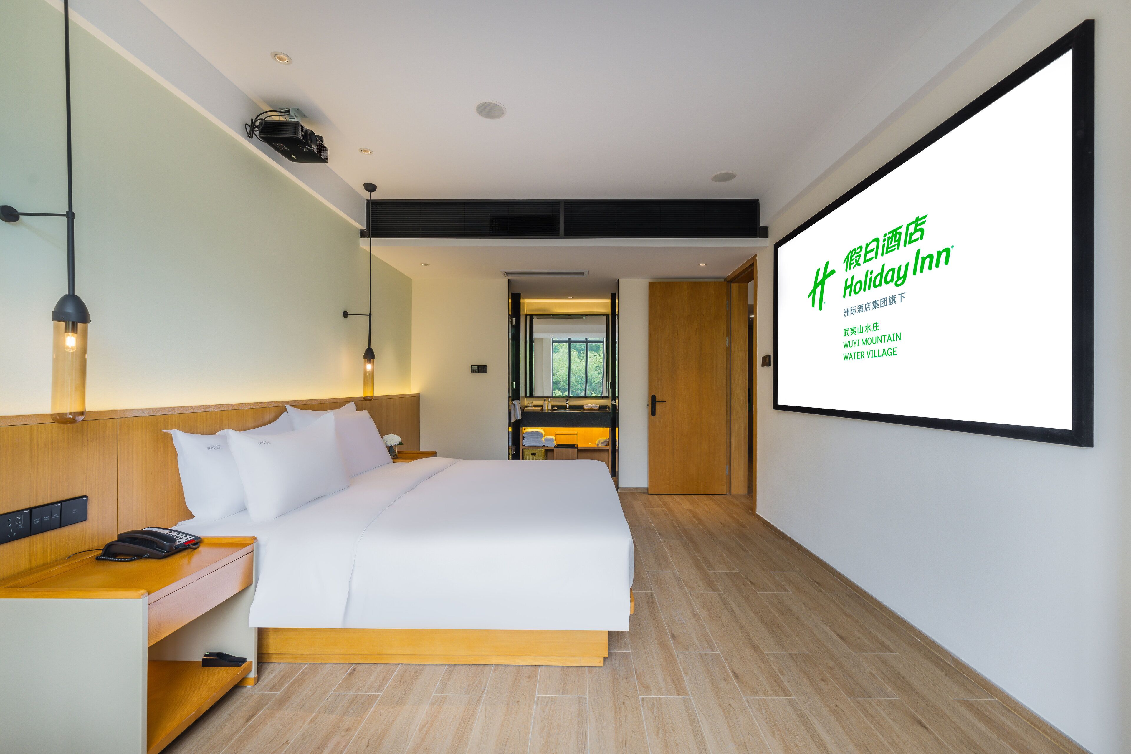 A modern hotel room with a large bed, bedside table, and a desk with a chair. A large screen displays the hotel name and location.