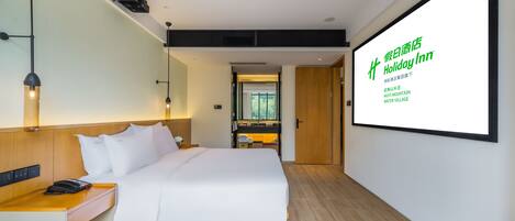 A modern hotel room with a large bed, bedside table, and a desk with a chair. A large screen displays the hotel name and location.