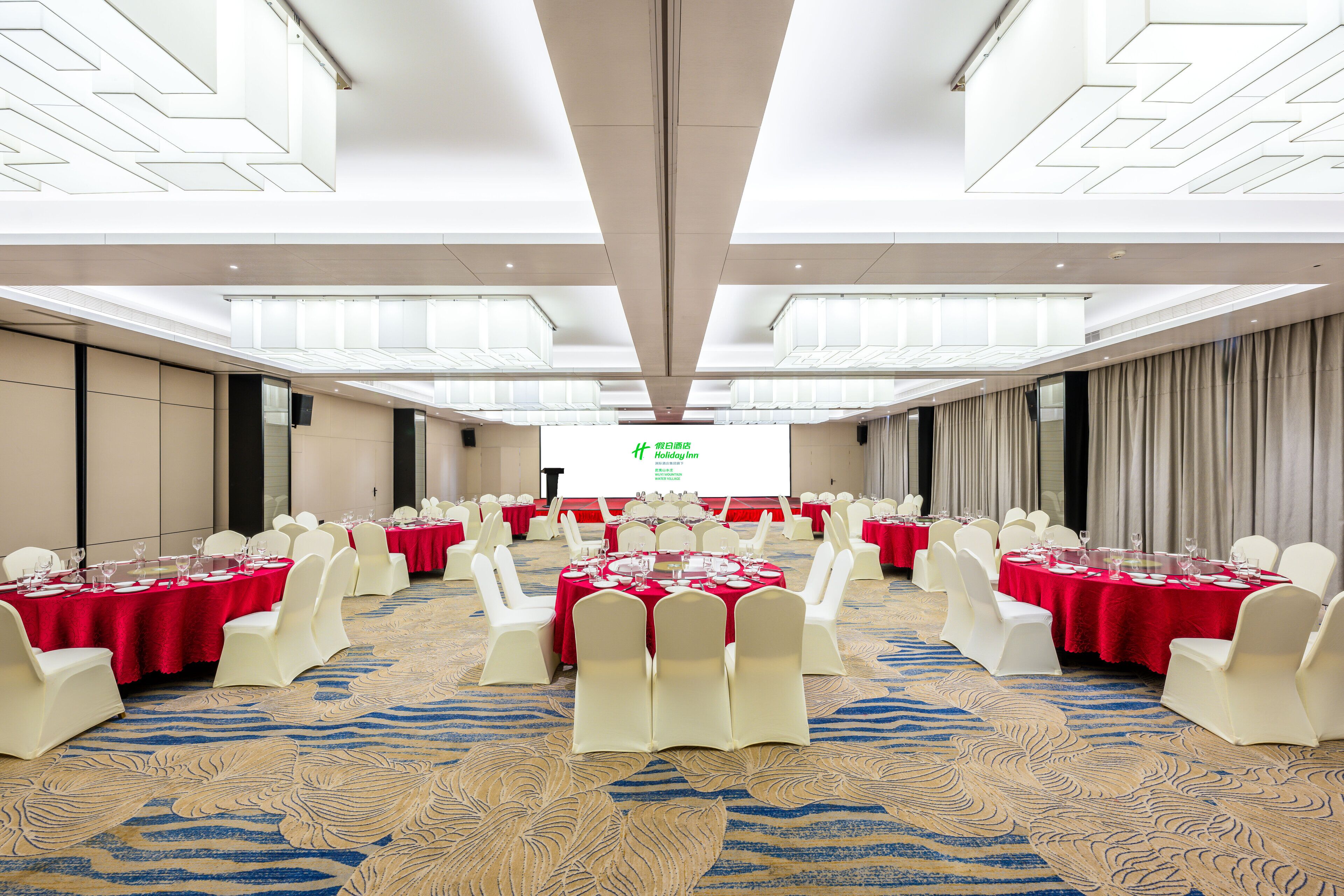 Un salón de banquetes con mesas redondas, sillas blancas y manteles rojos.