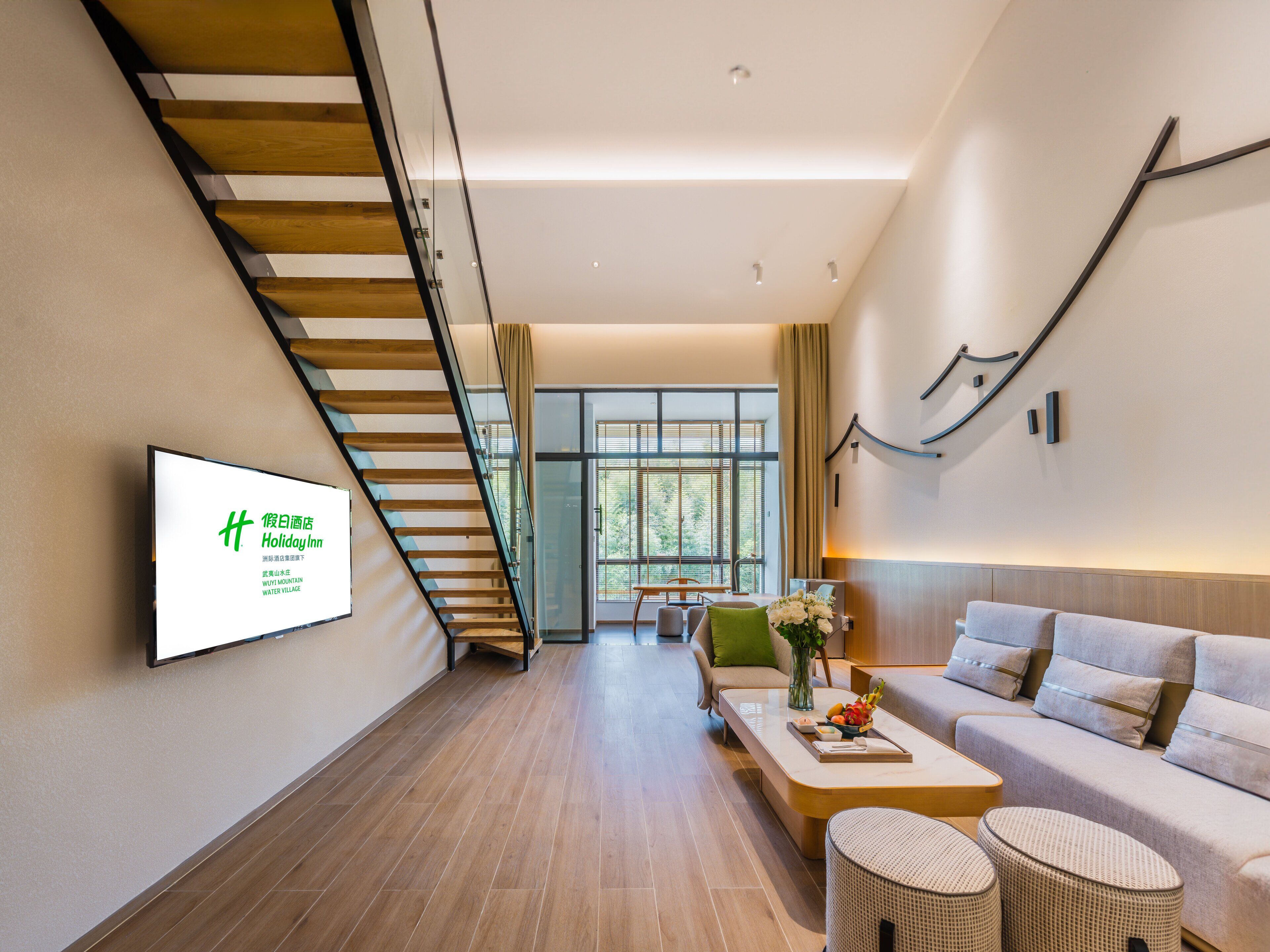 A modern hotel interior with a staircase, a living area with a sofa and coffee table, and a wall-mounted TV displaying 'Holiday Inn'.