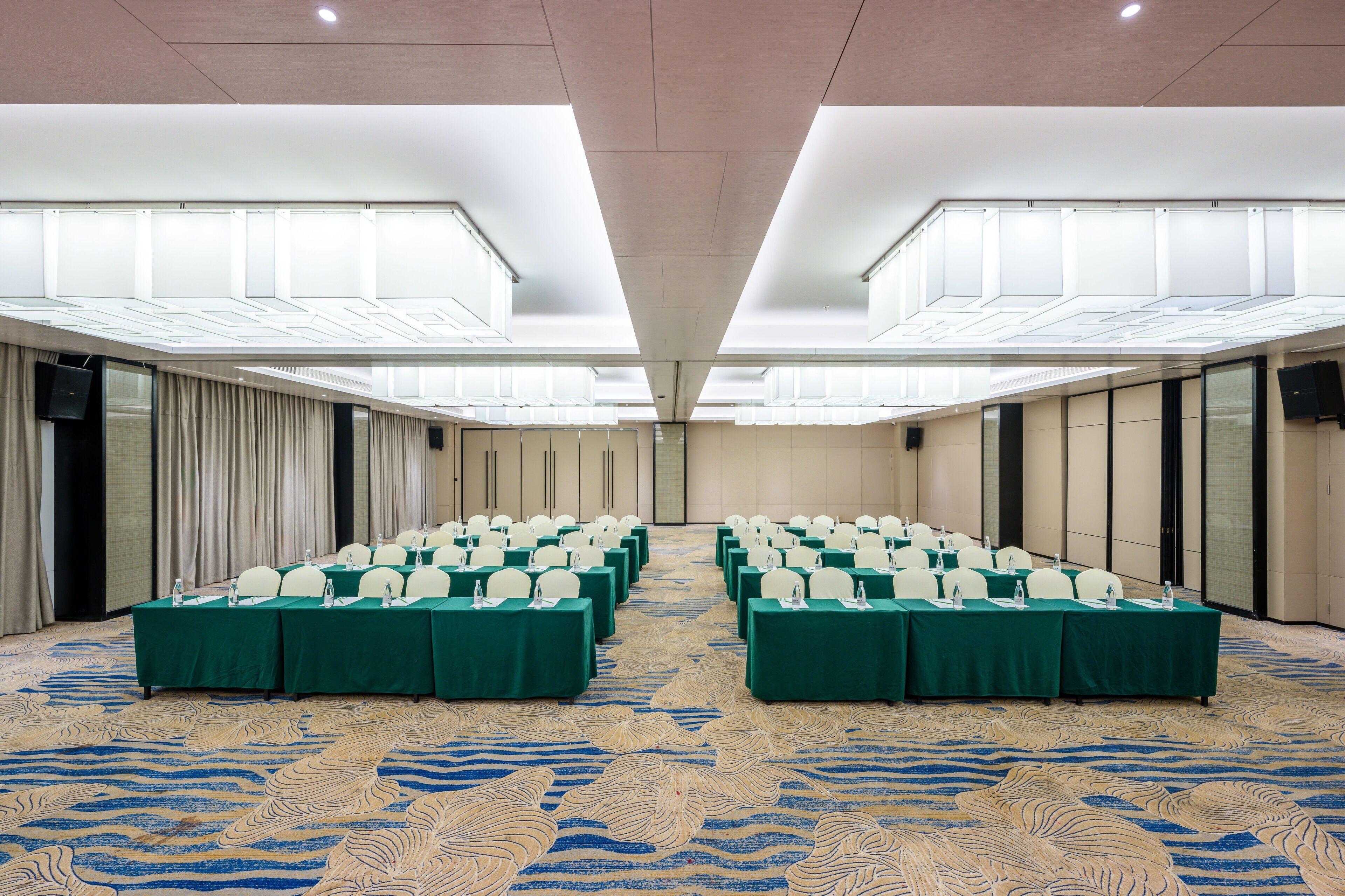 Un salón de conferencias con filas de mesas y sillas, una alfombra estampada y un techo alto con tragaluces.